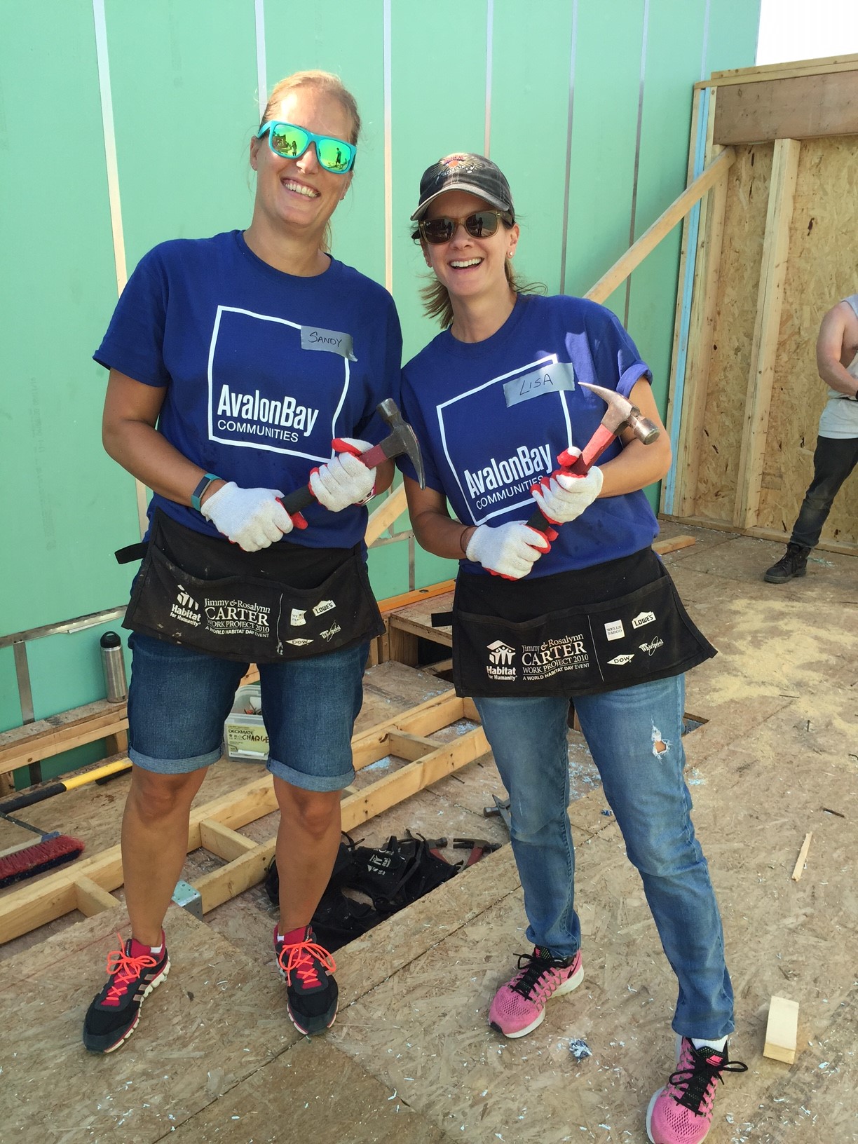 Two people smiling at the camera wearing blue t-shirts, sunglasses, and gloves holding hammers