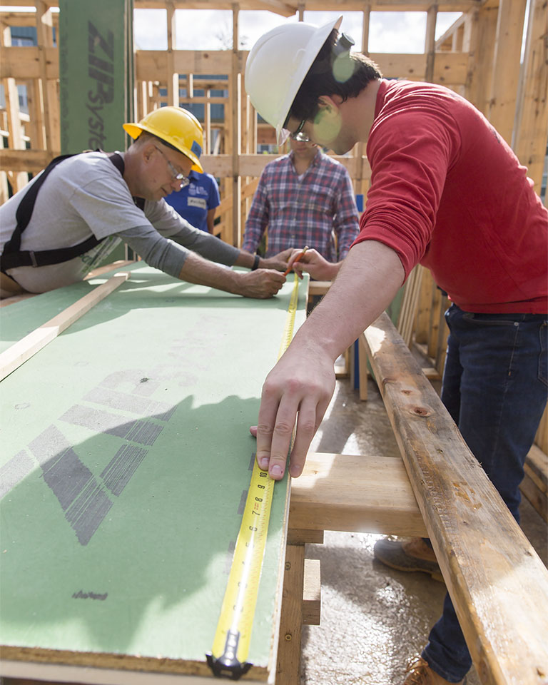 Three people at a construction site wearing headgear and using measuring tape on lengths of wood