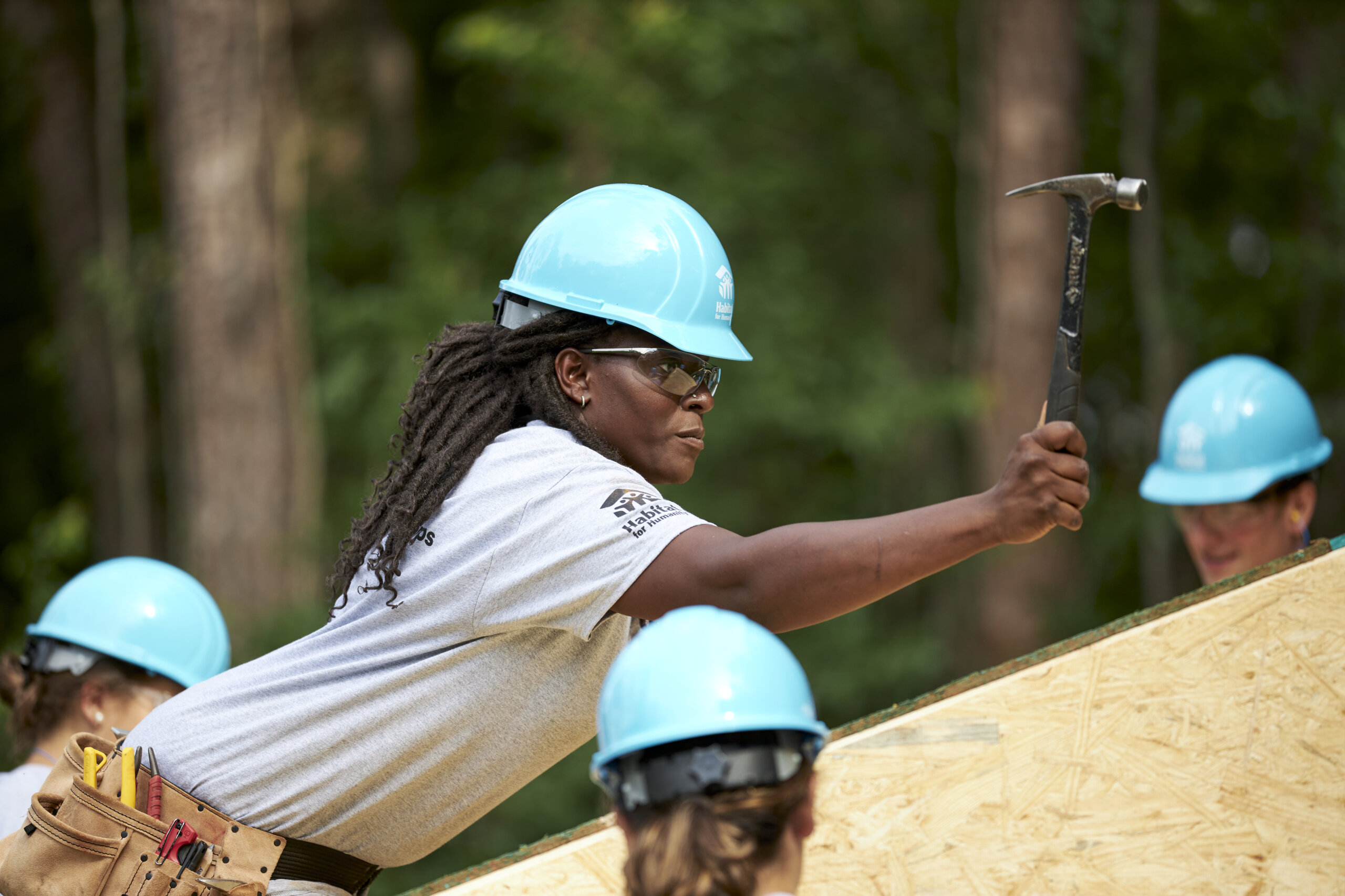 Woman leaning over and hammering a wall wearing a blue safety hardhat