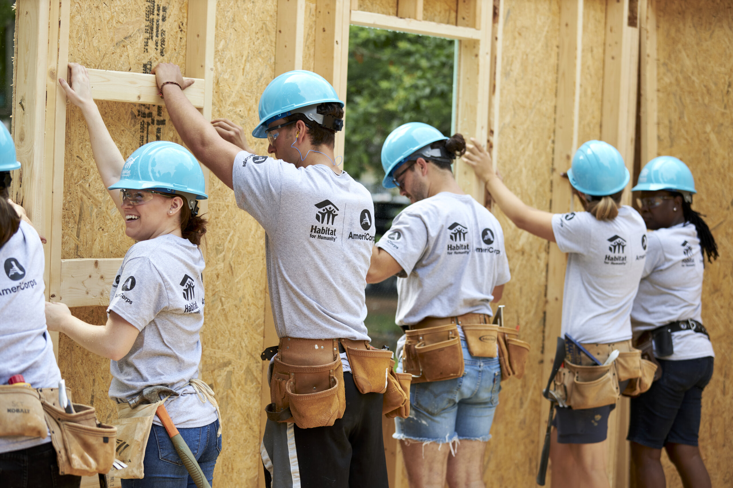 Group of people wearing blue hardhats and grey t-shirts that say "AmeriCorps" on back holding up a wall