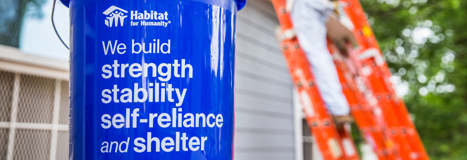 A blue sign that reads "we build strength, stability, self-reliance, and shelter" in front of a ladder
