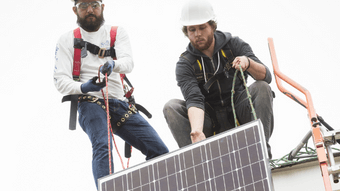 Two men wearing safety equipment installing a solar panel on a roof