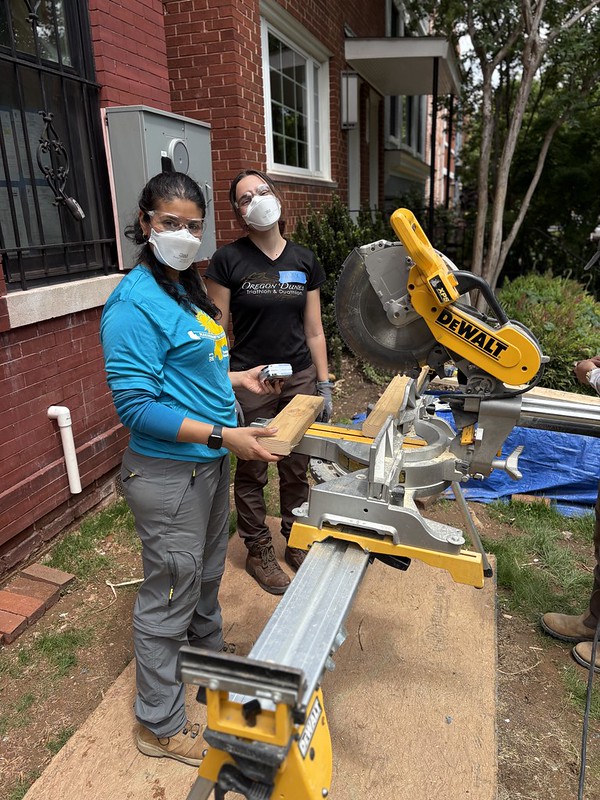 two women wearing masks and goggles, cutting wood pieces with a large tool